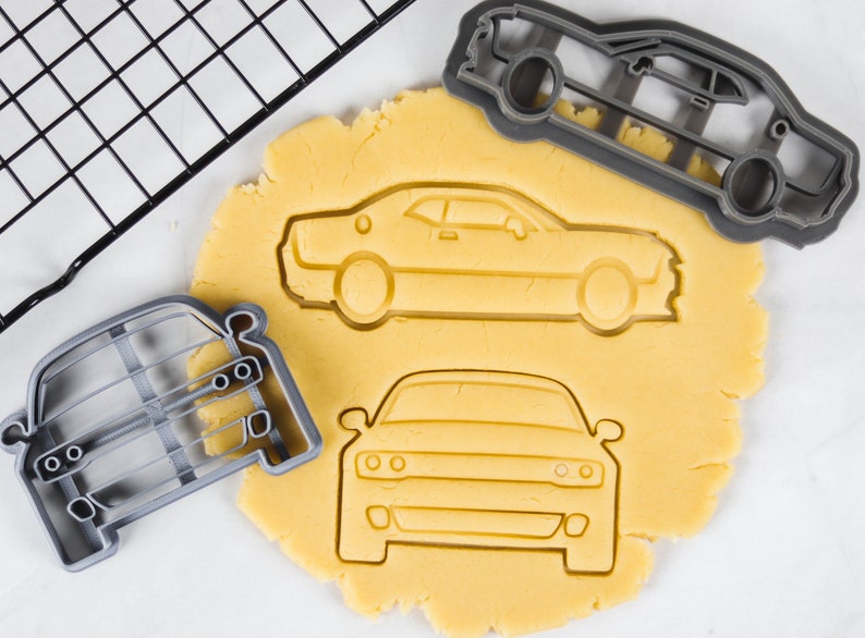 May include: Three cookie cutters in the shape of a car. The cutters are made of gray plastic and are on a white surface. The cutters are next to a sheet of yellow cookie dough.