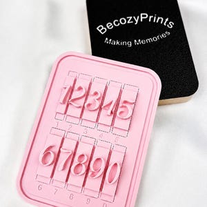 May include: A pink number stamp featuring the digits 1 to 0. The stamp has a rectangular shape with a raised edge. A black rectangular block with the text "BecozyPrints Making Memories" is in the background.