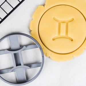 Op de afbeelding: Een grijze plastic koekjesvorm van het sterrenbeeld Tweelingen naast een cirkel van rauw koekjesdeeg met het Tweelingen-symbool erin gedrukt. Een zwart metalen afkoelrek staat in de linkerbovenhoek. De koekjesvorm is rond met het Tweelingen-symbool in het midden.