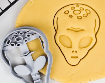 May include: A gray plastic cookie cutter in the shape of an alien head. The cutter is on top of a yellow cookie dough. The alien has large eyes, a small mouth, and a bumpy head.