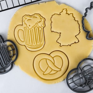 May include: A close-up of a cookie cutter set with a beer mug, pretzel, and Germany outline. The cookie cutters are black and the dough is a light yellow color.