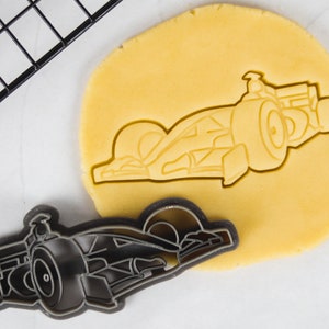 Pode incluir: Um cortador de biscoitos de plástico preto em forma de carro de corrida. O cortador de biscoitos está em cima de uma folha de massa de biscoitos amarela.