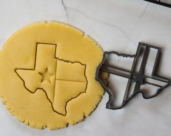 Könnte beinhalten: Eine Nahaufnahme eines Ausstechers in Form des Bundesstaates Texas mit einem Stern in der Mitte. Der Ausstecher liegt auf einer weißen Oberfläche neben einem Stück Keksteig, das mit dem Ausstecher ausgestochen wurde.