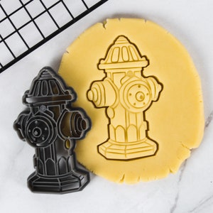 May include: A black metal cookie cutter shaped like a fire hydrant. The cookie cutter is on top of a yellow sugar cookie.