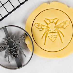 Könnte beinhalten: Ein Ausstecher in Form einer Biene. Der Ausstecher ist aus Metall und hat ein detailliertes Design. Der Ausstecher liegt auf einem Stück Keksteig.