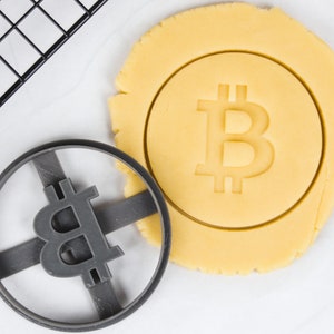 Könnte beinhalten: Eine Nahaufnahme eines Ausstechers in Form eines Bitcoin-Symbols. Der Ausstecher ist aus grauem Kunststoff und hat ein kreuzförmiges Design. Ein Keksteig wird mit dem Ausstecher ausgestochen.