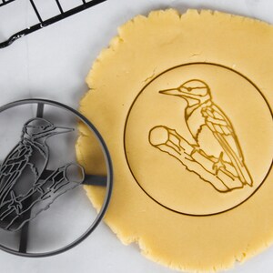 Può includere: Un tagliapasta in metallo nero a forma di picchio posato su un ramo. Il tagliapasta viene utilizzato per tagliare un biscotto da un foglio di pasta per biscotti non cotta.