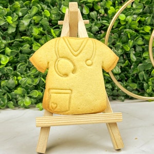 May include: A yellow sugar cookie shaped like a doctor's scrubs with a stethoscope and a pocket. The cookie is displayed on a small wooden easel.