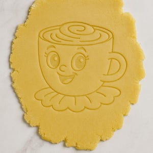 Könnte beinhalten: Ausstecher in Form einer lächelnden Kaffeetasse mit Rüschenkragen, in ein gelbes Teigblatt gedrückt. Die Tasse hat große Augen und eine Wirbel aus Schlagsahne oben. Der Teig liegt auf einer weißen Oberfläche.