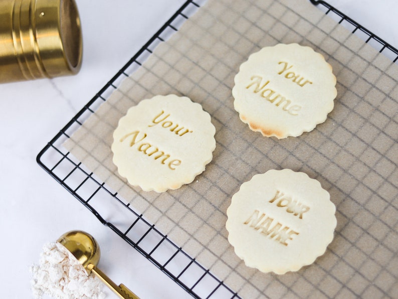 May include: Three plain sugar cookies on a wire rack with parchment paper. Each cookie has the words "Your Name" imprinted on the top.