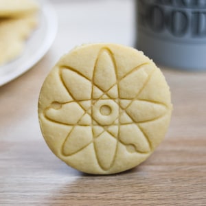 May include: A close-up of a single, round, light brown cookie with a detailed atom design embossed on the surface.