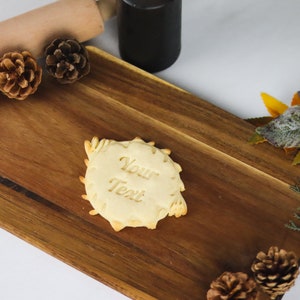 May include: A plain, round cookie with the words "Your Text" embossed on the surface. The cookie is on a wooden cutting board with pine cones and fall foliage around it.