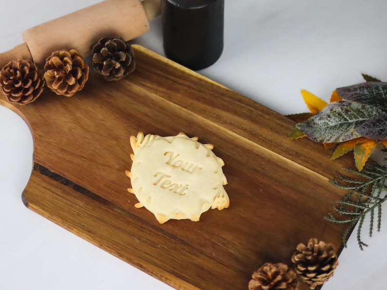 May include: A plain, round cookie with the words "Your Text" embossed on the surface. The cookie is on a wooden cutting board with pine cones and fall foliage around it.