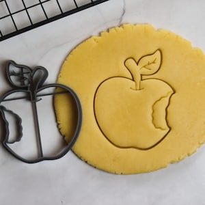 Peut inclure: Un emporte-pièce en métal gris en forme de pomme avec une bouchée, à côté d'un cercle de pâte jaune avec la forme de pomme pressée. Une grille de refroidissement noire est en arrière-plan.