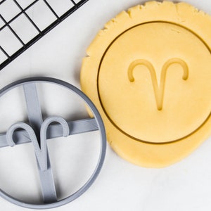 Puede incluir: Un cortador de galletas de plástico gris en forma del símbolo astrológico de Aries. El cortador de galletas está sobre una superficie blanca junto a una galleta con el mismo símbolo impreso en ella.