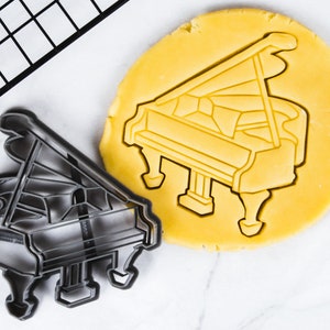 Pode incluir: Um cortador de biscoitos de metal preto em forma de piano de cauda. O cortador de biscoitos está sobre uma superfície branca ao lado de um recorte de massa de biscoito amarela em forma de piano de cauda.