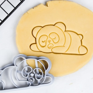 Puede incluir: Un cortador de galletas de plástico gris en forma de panda durmiendo. El cortador está sobre una hoja de masa para galletas sin hornear.