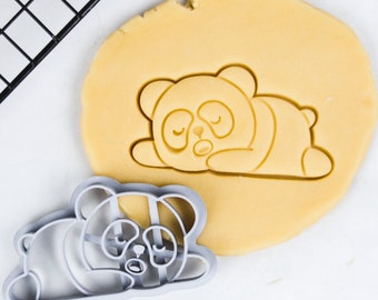 May include: A gray plastic cookie cutter in the shape of a sleeping panda bear. The cutter is on top of a sheet of unbaked cookie dough.