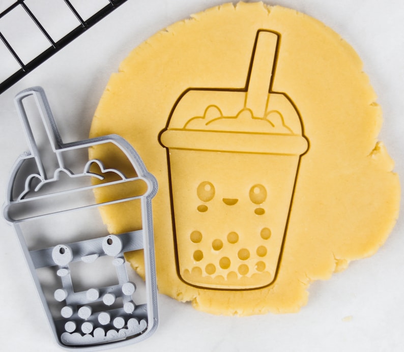 May include: A grey plastic cookie cutter shaped like a boba tea cup with a straw and a cute cartoon face. The cutter is on top of a yellow cookie dough.