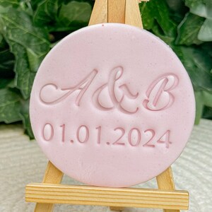 Peut inclure: Une plaque circulaire rose avec le texte "A&B 01.01.2024" en écriture cursive, présentée sur un petit chevalet en bois.