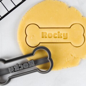 Op de afbeelding: Een grijze plastic koekjesvormer in de vorm van een hondenbot met het woord "Rocky" erop gegraveerd. De vormer ligt op een geel koekdeeg met dezelfde vorm en hetzelfde woord uitgesneden.