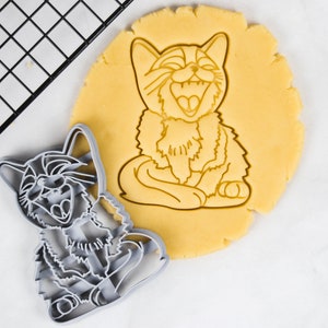 Può includere: Un tagliapasta in metallo grigio a forma di gatto con la bocca aperta in un'espressione di risata. Il tagliapasta è sopra un impasto per biscotti marrone chiaro con lo stesso disegno di gatto tagliato.