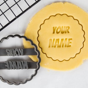 May include: A black plastic cookie cutter with the words "YOUR NAME" in a bold font. The cutter is on top of a round piece of unbaked cookie dough with the same words imprinted on it.