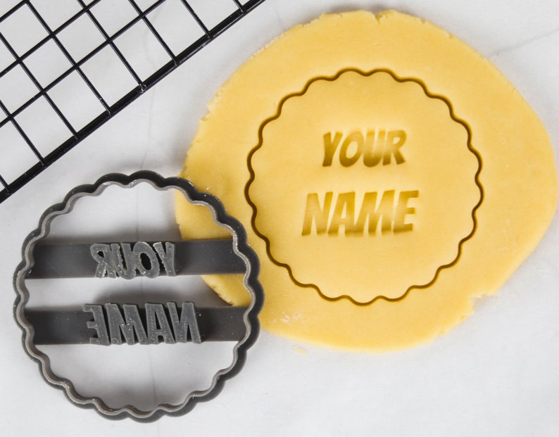 May include: A black plastic cookie cutter with the words "YOUR NAME" in a bold font. The cutter is on top of a round piece of unbaked cookie dough with the same words imprinted on it.
