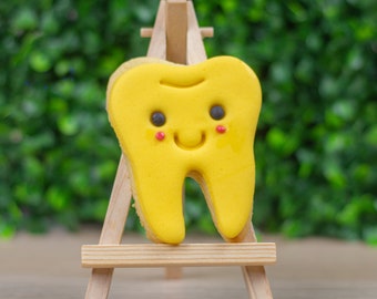 May include: A yellow tooth-shaped cookie with a smiling face, decorated with red dots for cheeks, is displayed on a small wooden easel.