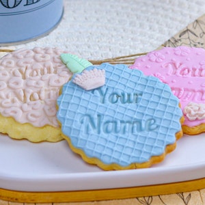 May include: Three decorated sugar cookies on a white oval plate. The cookies are decorated with pink, blue, and white icing. The text "Your Name" is written on the blue cookie. The text "Your Name" is written on the pink cookie.