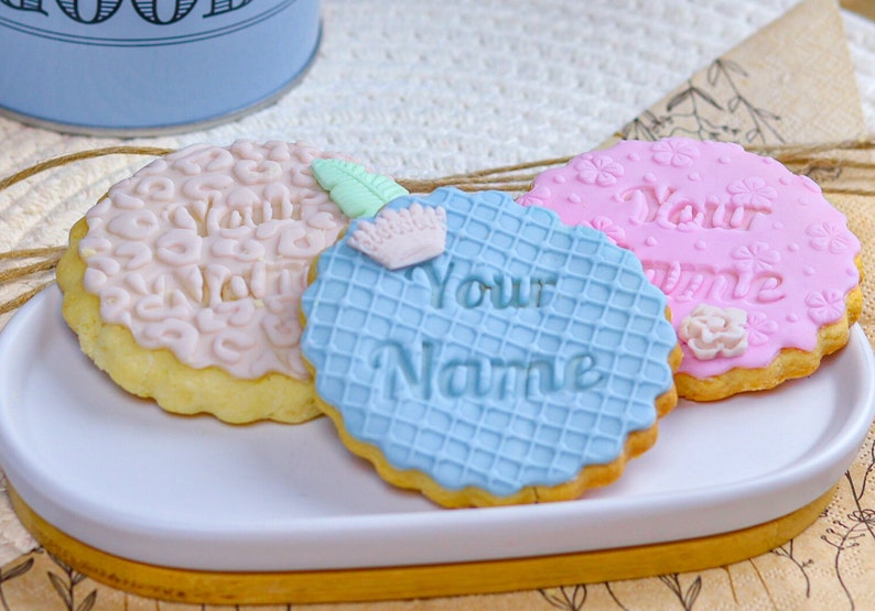 May include: Three decorated sugar cookies on a white oval plate. The cookies are decorated with pink, blue, and white icing. The text "Your Name" is written on the blue cookie. The text "Your Name" is written on the pink cookie.