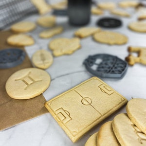 May include: A close-up of a variety of homemade sugar cookies, including a football pitch, a zodiac sign, and a stack of plain cookies. The cookies are on a white marble surface.