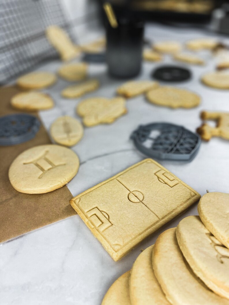 May include: A close-up of a variety of homemade sugar cookies, including a football pitch, a zodiac sign, and a stack of plain cookies. The cookies are on a white marble surface.
