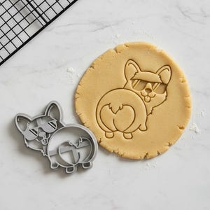 May include: A gray corgi cookie cutter next to a round of dough with a corgi imprint. The corgi design features sunglasses. A black cooling rack is in the background. The dough is light brown.