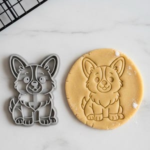 May include: A gray plastic corgi dog cookie cutter next to a round cookie with a corgi dog imprint. The cookie cutter is a detailed outline of the dog. The cookie is light brown and has the dog's features pressed into it.