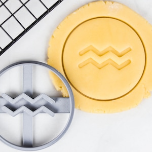 May include: A gray plastic cookie cutter with the Aquarius symbol next to a piece of dough with the same imprint. The dough is yellow and the cutter is gray. A black cooling rack is in the background.
