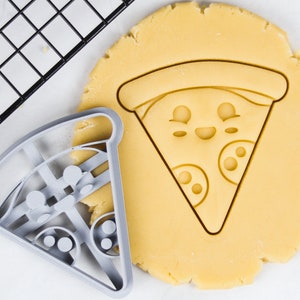 May include: A grey plastic cookie cutter shaped like a pizza slice with a smiling face and pepperoni. The cutter is on top of a round piece of unbaked cookie dough.
