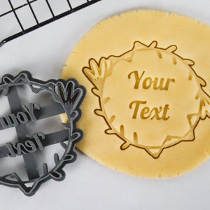 May include: A gray plastic cookie cutter in the shape of a wreath with the words "Your Text" inside. A plain cookie dough is shown with the cutter pressed into it.