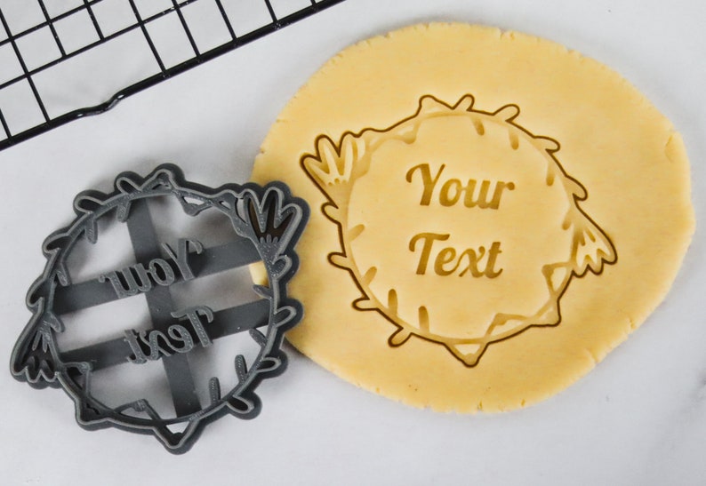 May include: A gray plastic cookie cutter in the shape of a wreath with the words "Your Text" inside. A plain cookie dough is shown with the cutter pressed into it.