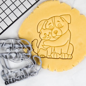 May include: A gray plastic cookie cutter in the shape of a dog hugging a cat. The cookie cutter is on top of a round piece of unbaked cookie dough.