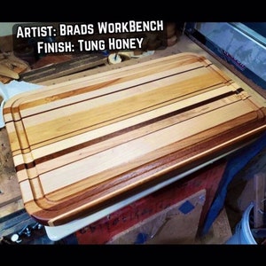 May include: A large, rectangular wooden cutting board with a rounded edge. The board is made of multiple types of wood, creating a striped pattern. The board is finished with a Tung Honey finish. The text "ARTIST: BRADS WORKBENCH FINISH: TUNG HONEY" is visible at the top of the image.