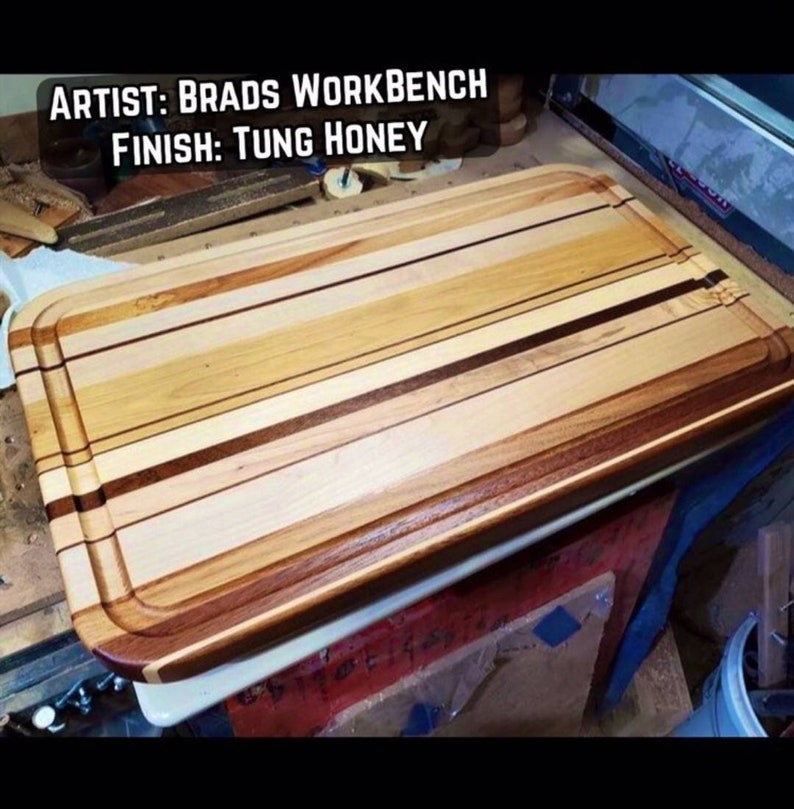 May include: A large, rectangular wooden cutting board with a rounded edge. The board is made of multiple types of wood, creating a striped pattern. The board is finished with a Tung Honey finish. The text "ARTIST: BRADS WORKBENCH FINISH: TUNG HONEY" is visible at the top of the image.