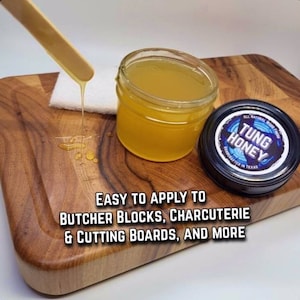 May include: A clear glass jar filled with a yellow liquid, labeled "TUNG HONEY" with a blue and white logo. The jar is sitting on a wooden cutting board with a wooden stick dripping the liquid onto the board. The text "EASY TO APPLY TO BUTCHER BLOCKS, CHARCUTERIE & CUTTING BOARDS, AND MORE" is written below the jar.