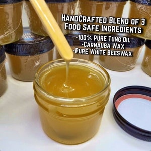May include: A close-up of a clear glass jar filled with a golden yellow substance. A wooden stick is dipping into the jar, and the substance is dripping from the stick. The jar is labeled "HANDCRAFTED BLEND OF 3 FOOD SAFE INGREDIENTS - 100% PURE TUNG OIL - CARNAUBA WAX - PURE WHITE BEESWAX".
