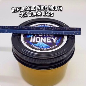 May include: A 4 ounce glass jar with a wide mouth, filled with honey. The jar has a black lid with a label that says "YUM HONEY" in blue and white text. The label also says "HANDCRAFTED IN TEXAS".