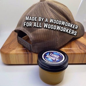 May include: A brown and tan mesh trucker hat with the text "MADE BY A WOODWORKER FOR ALL WOODWORKERS" on the back. The hat is sitting on a wooden cutting board with a small jar of "TUNG HONEY" on the board.
