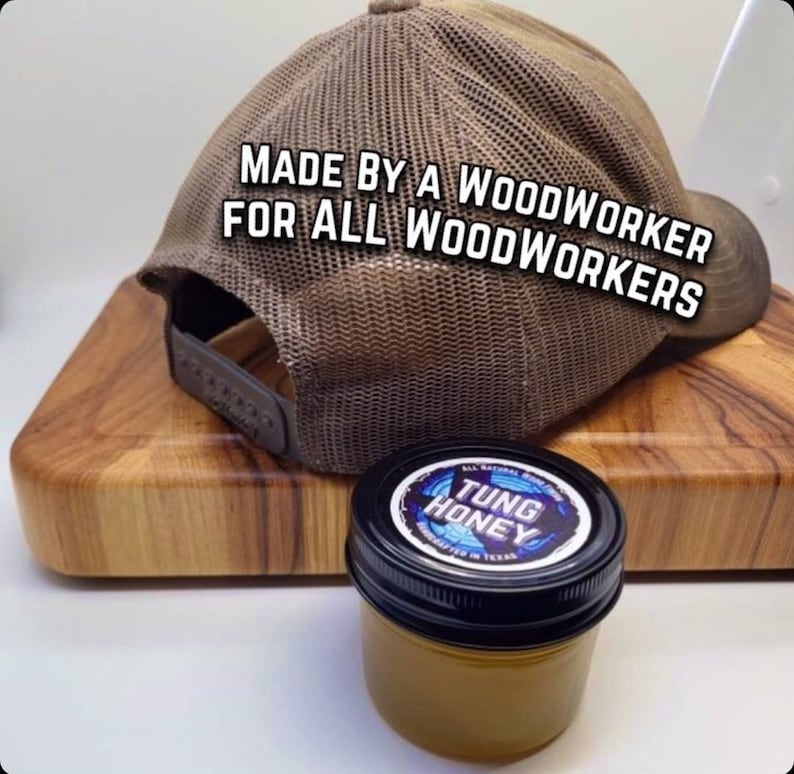 May include: A brown and tan mesh trucker hat with the text "MADE BY A WOODWORKER FOR ALL WOODWORKERS" on the back. The hat is sitting on a wooden cutting board with a small jar of "TUNG HONEY" on the board.