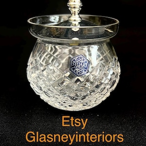 Peut inclure: Un pot en cristal transparent avec un motif taillé en diamant et un couvercle argenté. Le pot a une forme ronde et une petite poignée décorative sur le dessus. Le pot porte une étiquette bleue et blanche avec "STUART CRYSTAL". Le texte "Etsy Glasneyinteriors" est en orange.