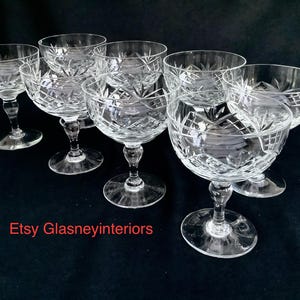 May include: A collection of six clear crystal champagne coupe glasses. Each glass has a round bowl with a cut crystal pattern and a stem. The glasses are displayed on a dark background. The text "Etsy Glasneyinteriors" is visible.