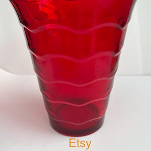 May include: A bright red glass vase with a wavy rim and horizontal wave-like ridges. The vase has a flared top and a cylindrical body. The Etsy shop name "Glasneyinteriors" is printed at the bottom.